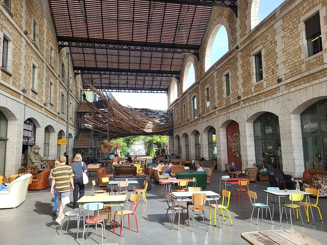 Sous la halle de Darwin à Bordeaux, des tables et des fauteuils dépareillés devant la façade en pierre du Magasin Général.