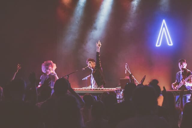 Un chanteur-pianiste bras levé sur scène, entouré de deux musiciens, face à un public dans la pénombre sous des faisceaux de lumière