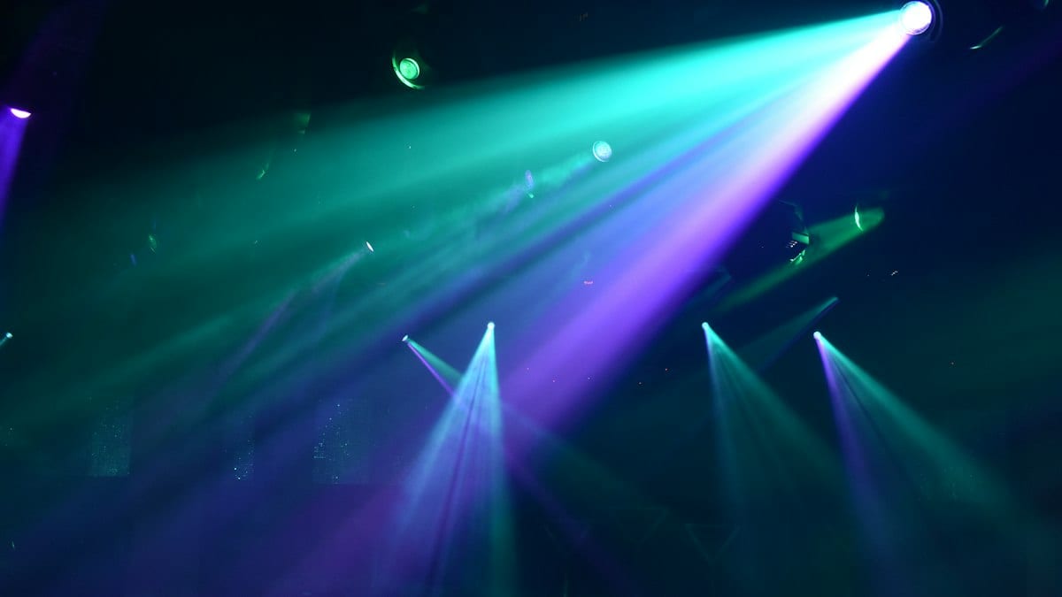 Faisceaux de projecteurs verts et violets traversant la fumée d'une salle de concert plongée dans le noir.
