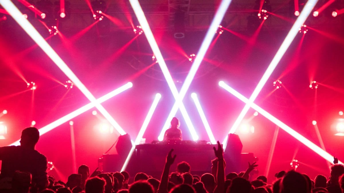 Foule les bras levés devant un DJ, sous des faisceaux lumineux blancs et roses croisés dans une salle de concert.