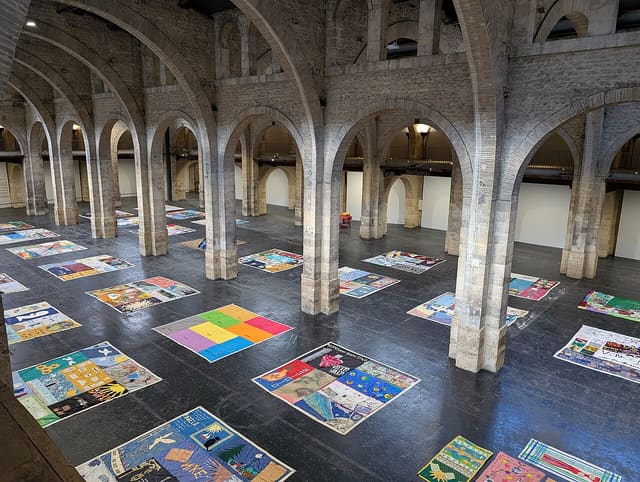 Vue en hauteur de la nef de l'Entrepôt Lainé à Bordeaux, avec ses arcades de pierre et des œuvres textiles posées au sol.